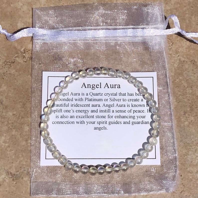 May include: A white Angel Aura bracelet made of clear quartz crystals with iridescent rainbow hues. The bracelet is displayed on a white organza bag with a card that reads "Angel Aura is a Quartz crystal that has been bonded with Platinum or Silver to create a beautiful iridescent aura. Angel Aura is known to uplift one's energy and instill a sense of peace. It is also an excellent stone for enhancing your connection with your spirit guides and guardian angels."