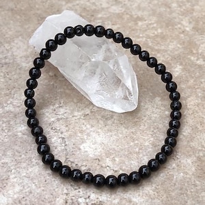 May include: A black beaded bracelet made of small, round beads. The bracelet is lying on a white, textured surface.