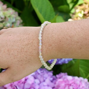 May include: A delicate bracelet made of iridescent white beads. The bracelet is worn on a person's wrist, with a blurred background of purple flowers.