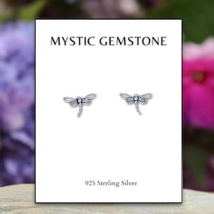 May include: A pair of sterling silver dragonfly stud earrings on a white card. The card reads "MYSTIC GEMSTONE" at the top and "925 Sterling Silver" at the bottom. The earrings are detailed with delicate wings and a slender body.
