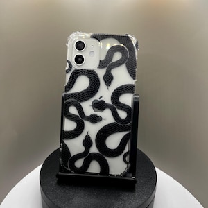 May include: Transparent phone case featuring a black snake design. The case, designed for a smartphone, has a repeating pattern of black snakes against a clear background. The phone case is displayed on a black stand.