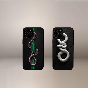 May include: Two black phone cases with snake designs. One case features a green stripe with a red and silver snake. The other case has a white snake design. Both cases have a textured appearance.