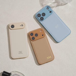 May include: Three personalised phone cases in cream, tan, and light blue. Each case has a different colour camera lens surround and initials. A small perfume bottle with a black cap and a glass are also in the image.