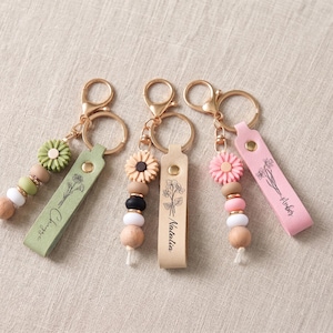 May include: Three keychains featuring gold-tone lobster clasps and rings. Each has a leather-like strap with a printed floral design and a name: "Chayse" on sage green, "Natalia" on beige, and "Amber" on pink. Each includes a flower-shaped bead.