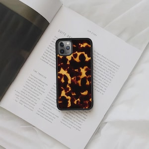 Op de afbeelding: Een smartphonehoesje met een schildpadpatroon. De hoes heeft zwarte randen en een herhalend patroon van bruine en gele vormen. Het telefoonhoesje ligt op een open boek.
