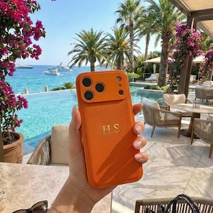 Könnte beinhalten: Orangefarbene Handyhülle mit goldenen Initialen "H.S.". Die Hülle wird vor einem Hintergrund mit Pool, Meer, Palmen und Booten gehalten. Sie hat einen Kameraausschnitt.