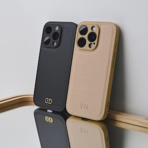 Puede incluir: Dos fundas para smartphone con acabado texturizado, una en negro con las iniciales GD en dorado y otra en beige con las iniciales YN en dorado. Ambas fundas cuentan con un recorte elevado para la cámara y se reflejan en una superficie lisa.
