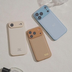 May include: Three rectangular phone cases in beige, tan, and light blue. Each case features a camera cutout area in a matching or contrasting tone and is personalised with two-letter initials: JK on the beige case, NM on the tan case, and SP on the blue case.