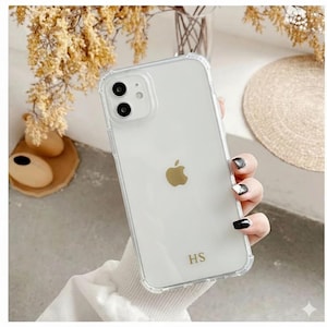 May include: Clear iPhone case with a gold Apple logo and the initials "HS" in gold. The case has a protective bumper around the edges. The phone is held in a hand with manicured nails.