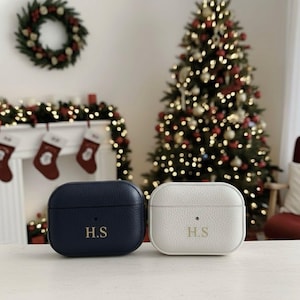 May include: Two personalized AirPod cases, one navy blue and one white, each with the initials "H.S." in gold lettering. The cases are displayed on a white surface, with a Christmas tree and decorations in the background.
