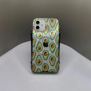 May include: A clear phone case with a pattern of green avocados on a white background. The case has a ring holder on the back. The text "NESOYAM" is printed on the bottom of the case.
