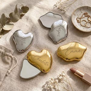 May include: Four irregular, organic-shaped pocket mirrors with metallic finishes. Two are silver-toned, engraved with the names Mia and Harper, and two are gold-toned, engraved with the names Charlotte and Emma. Each mirror features a polished, reflective surface.