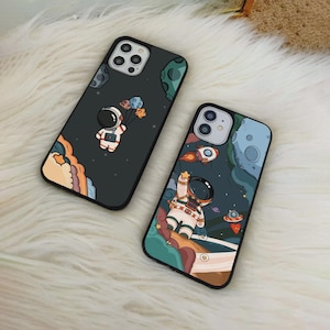 Puede incluir: Dos fundas para teléfono con diseños espaciales. Una funda presenta un astronauta sosteniendo globos contra un fondo oscuro con planetas. La otra funda muestra a un astronauta alcanzando una estrella con un cohete y un OVNI. Ambas fundas tienen un borde negro.