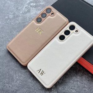 Pode incluir: Duas capas de telefone personalizadas. Uma é rosa claro com as iniciais "HK" em dourado. A outra é branca com as iniciais "AW" em preto. Ambas as capas têm uma superfície texturizada e um módulo de câmera.