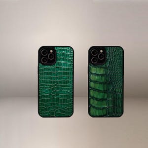 Op de afbeelding: Twee groene telefoonhoesjes met een krokodillenleertextuur. De hoesjes hebben een zwarte rand en een camera-uitsparing. De hoesjes zijn verticaal georiënteerd.