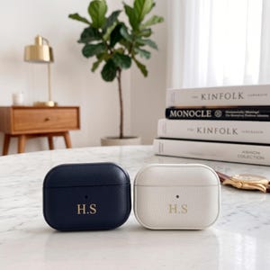 May include: Two textured leather earbud cases, one navy blue and one white, each featuring the gold embossed initials H.S on the front. The cases have a rounded rectangular shape with a small circular cutout for a charging indicator light.