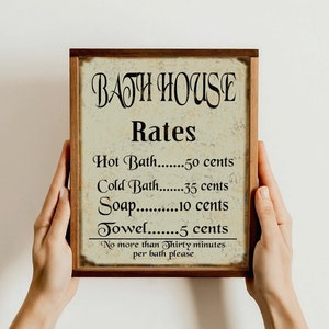 Puede incluir: Un cartel de estilo vintage con marco de madera y fondo blanco. El cartel dice "BATH HOUSE Rates" y enumera los precios de los ba&ntilde;os calientes, los ba&ntilde;os fr&iacute;os, el jab&oacute;n y las toallas. El texto es negro y el cartel tiene un aspecto desgastado.