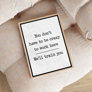 May include: A framed print with the text "You don't have to be crazy to work here. We'll train you." The print is sitting on a light brown couch.