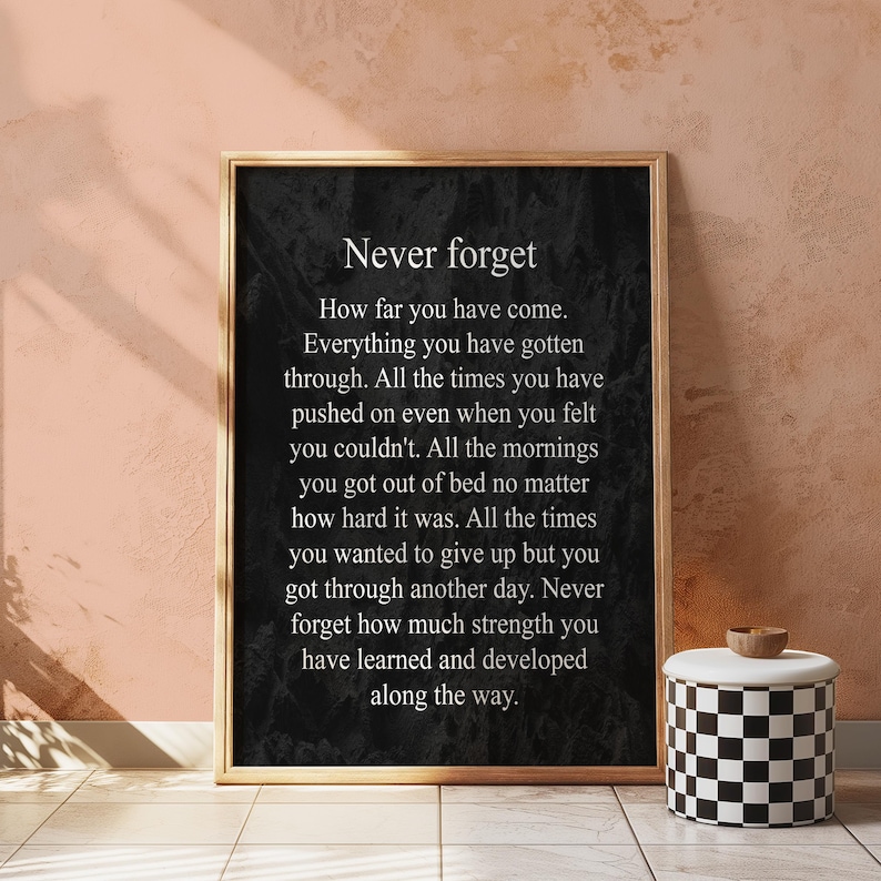 May include: Black and white inspirational quote poster with the text "Never forget" and a message about perseverance and strength. The poster is framed in a gold frame and is hanging on a wall.