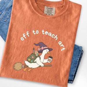 May include: A burnt orange t-shirt featuring a graphic of a white goose wearing a purple witch hat, carrying a green backpack with art supplies, and holding a coffee cup while flying on a broomstick with the text off to teach art.