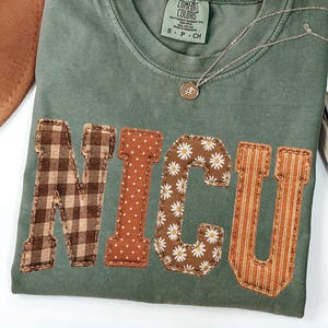 May include: A moss green short-sleeve t-shirt featuring the letters NICU appliqued in four distinct fabric patterns: brown gingham, brown with white polka dots, brown with white daisies, and orange with vertical stripes, with a tag reading Comfort Colors.
