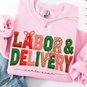 May include: A light pink crewneck sweatshirt featuring the text LABOR & DELIVERY in a patchwork style with red, green, and white patterns, accented by pink bows. Below the text, the name nurse anne is written in a black cursive script.