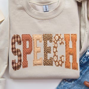May include: A beige crewneck sweatshirt featuring the word SPEECH in large, block-style letters across the chest. Each letter has a unique pattern, including brown plaid, polka dots, tan gingham, leopard print, white daisies, and orange stripes.