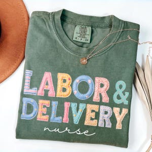 May include: A green Comfort Colors t-shirt with the words "LABOR & DELIVERY nurse" in colorful, patchwork-style letters. A gold necklace with a pendant rests on the shirt. A brown hat is partially visible in the upper left corner.
