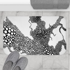 May include: A white bath mat with a black abstract design of interconnected shapes filled with varying sizes of black dots. The mat is rectangular and sits on a tiled floor. Gray slippers are at the bottom.