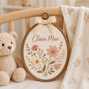 Puede incluir: Un bastidor de bordado ovalado de madera que contiene tela blanca con el nombre Clara Mae bordado en letra marrón, rodeado por un arreglo floral de flores rosas y crema con hojas verdes, rematado con un lazo de encaje color crema.