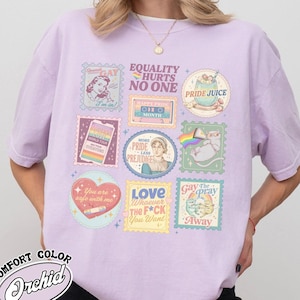 Puede incluir: Una camiseta de color lila claro con varios gráficos coloridos en forma de sello. Los sellos presentan frases como "Equality Hurts No One", "Pride Juice" y "Love Whoever The F*ck You Want". La camiseta también tiene el texto "Comfort Color Orchid".