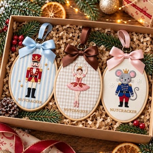 以下が含まれることがあります： 箱に入った3つの楕円形の刺繍枠。それぞれくるみ割り人形をテーマにしたデザインで、青いストライプの兵隊、ベージュのギンガムチェックのバレリーナ、制服を着たネズミが描かれています。それぞれにサテンのリボンとNUTCRACKERの文字が添えられています。