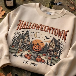 May include: A beige crewneck sweatshirt featuring a graphic print with the text HALLOWEENTOWN EST. 1998. The illustration depicts two houses, a crescent moon, a large orange pumpkin on a pedestal, smaller pumpkins, and black cat silhouettes.