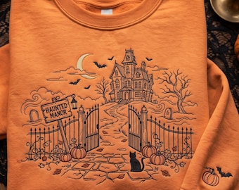 May include: An orange crewneck sweatshirt featuring a black embroidered design of a haunted manor, a crescent moon, bats, pumpkins, and a cat. The text "HAUNTED MANOR" is on a sign near the gate. A small pumpkin and bat are embroidered on the sleeve.