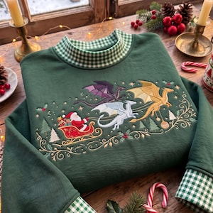May include: A dark green sweatshirt featuring a high collar and cuffs with a white and green gingham pattern. The chest displays an embroidered scene of Santa Claus in a red sleigh pulled by three dragons in purple, white, and gold colours.