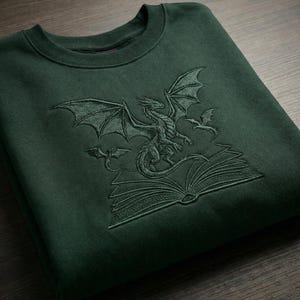 以下が含まれることがあります： 刺繍されたドラゴンデザインのダークグリーンスウェットシャツ。デザインには、開いた本にとまっているドラゴンと、2匹の小さなドラゴンが含まれています。スウェットシャツは柔らかい素材で作られています。