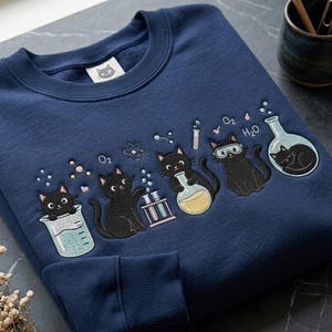 Peut inclure: Un sweat-shirt bleu marine à col rond orné d'une broderie représentant des chats noirs interagissant avec de la verrerie de laboratoire, notamment des béchers, des éprouvettes et des fioles, avec des formules chimiques O2 et H2O et un logo chat.
