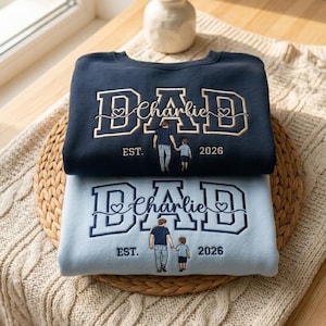May include: Two sweatshirts, one navy blue and one light blue, are stacked on a woven mat. The navy sweatshirt has "DAD" embroidered in gold with "Charlie" below. The light blue sweatshirt has the same design. Both have an illustration of a father and child.