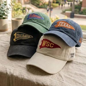 May include: Four baseball caps in black, olive green, blue, and cream. Each cap features a pennant-shaped patch with text: 'CHALHAVEN' in yellow, 'FLORMONT' in blue, 'TEXAL' in orange, and 'WSC' in red. The cream cap has 'GAME DAY' text on the side.