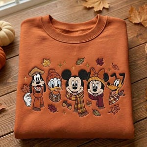 Peut inclure: Un sweat-shirt à col rond orange brûlé avec une broderie représentant Dingo, Donald, Mickey, Minnie et Pluto portant des écharpes et des bonnets d'hiver, entourés de petites feuilles d'automne et de glands brodés.