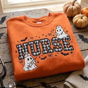 Può includere: Una felpa girocollo arancione con la scritta NURSE in lettere a quadri bianchi e neri. Il design include due fantasmi bianchi con cuffie da infermiere, uno che tiene una siringa e l'altro uno stetoscopio, oltre a piccoli pipistrelli.