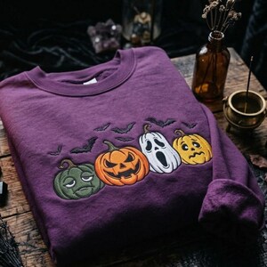May include: A deep purple crewneck sweatshirt featuring an embroidered design of four distinct pumpkins with varied facial expressions, including a green, orange, white, and yellow pumpkin, surrounded by small black embroidered bats.