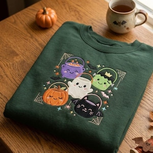 May include: A dark green crewneck sweatshirt featuring an embroidered design of five cute Halloween characters: a purple cauldron, a green Frankenstein monster, a white ghost, an orange pumpkin, and a black bat, all carrying small buckets.