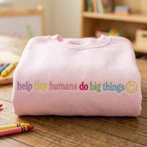 Puede incluir: Una sudadera de cuello redondo en color rosa claro con el texto bordado "help tiny humans do big things" en letras multicolores, seguido de un icono de cara sonriente bordado en amarillo en el centro de la prenda.