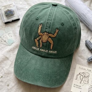 May include: A forest green baseball cap featuring an embroidered tan creature with multiple limbs and the text "AMAZE AMAZE AMAZE" centred on the front panel. The cap has a structured crown, a curved brim, and visible stitching details throughout.