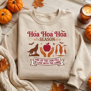 May include: A beige crewneck sweatshirt featuring a graphic print with the text "Hoa Hoa Hoa SEASON" and "AND SO THE LION FELL IN LOVE WITH THE LAMB". The design includes a wolf, a red apple held by two hands, and two white chess pieces.