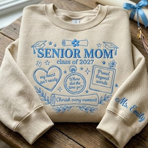 Pode incluir: Uma camisola bege de gola redonda com bordado azul claro com o texto SENIOR MOM class of 2027, my heart isn't ready, Where did the time go?, Proud beyond words, Cherish every moment e Ms. Emily no punho da manga.