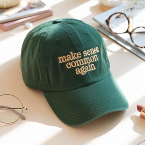 May include: A dark green baseball cap with the embroidered phrase "make sense common again" in a light tan color. The cap has a curved brim and a classic six-panel design. The hat is made of a soft fabric.