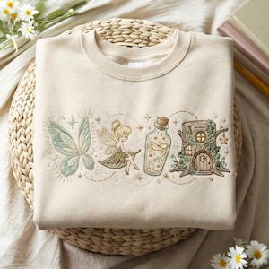 Puede incluir: Una sudadera de cuello redondo en color beige con bordados detallados en el pecho. El diseño incluye un par de alas de hada verde claro, una pequeña hada sentada sobre una hoja, una botella de cristal con polvo brillante y una casa en un tronco.
