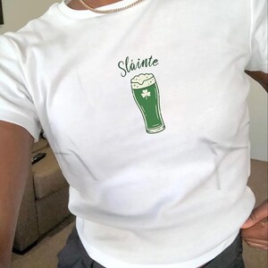 May include: White t-shirt with a green graphic of a pint of beer and the word "Sláinte" in a script font. The beer glass has a shamrock on it. The shirt is short-sleeved.